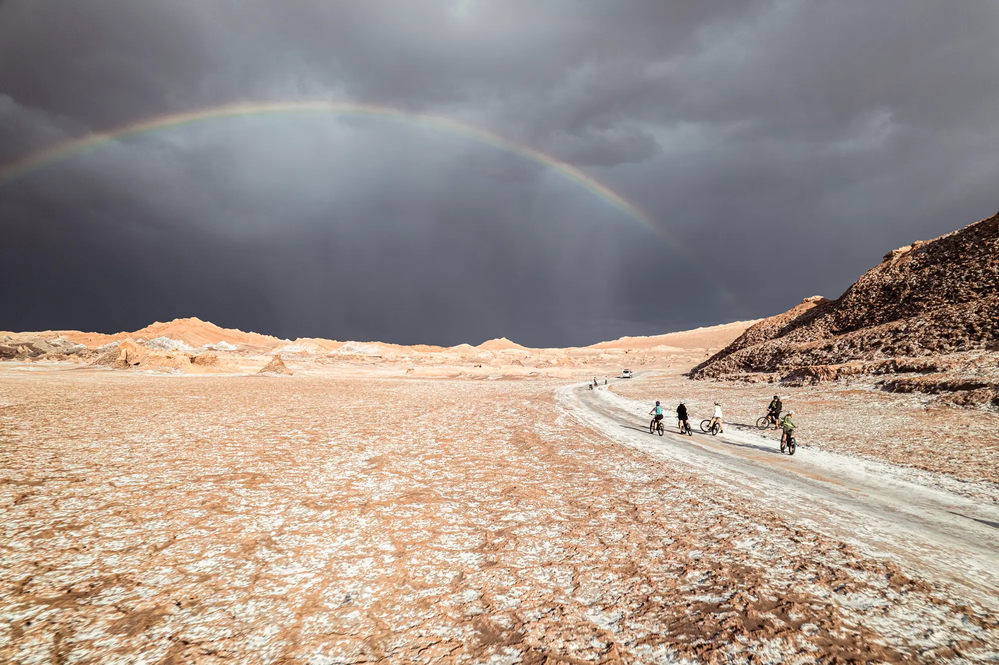 Cyclists crossing a salt plain under a rainbow in the Atacama